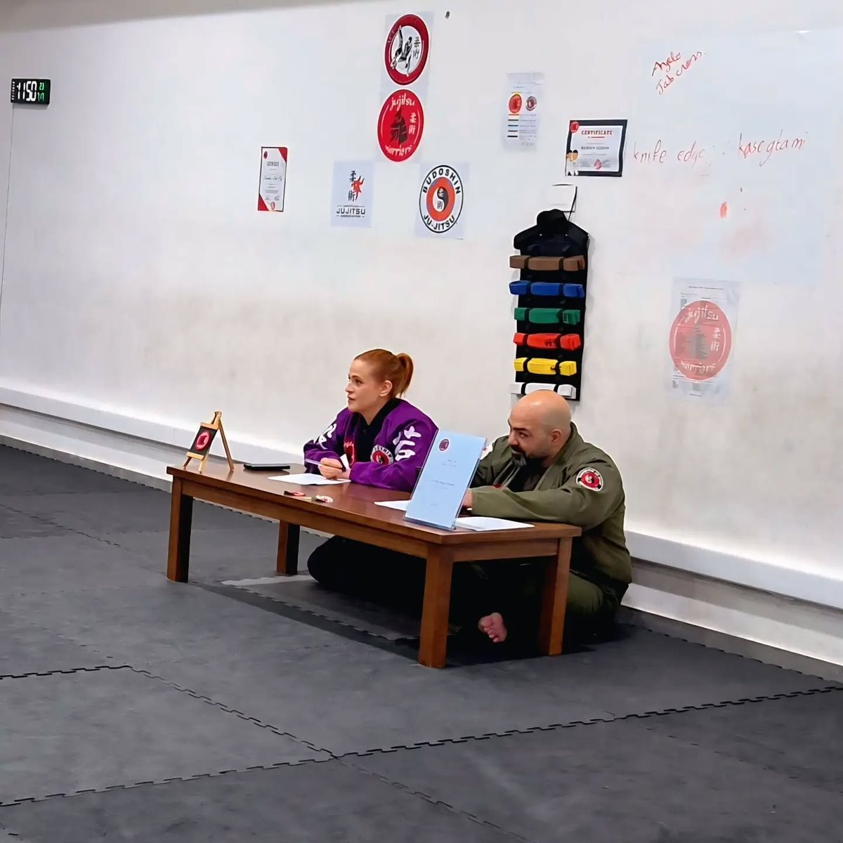 Two instructors seated at the grading table, our belt rack on the wall behind them